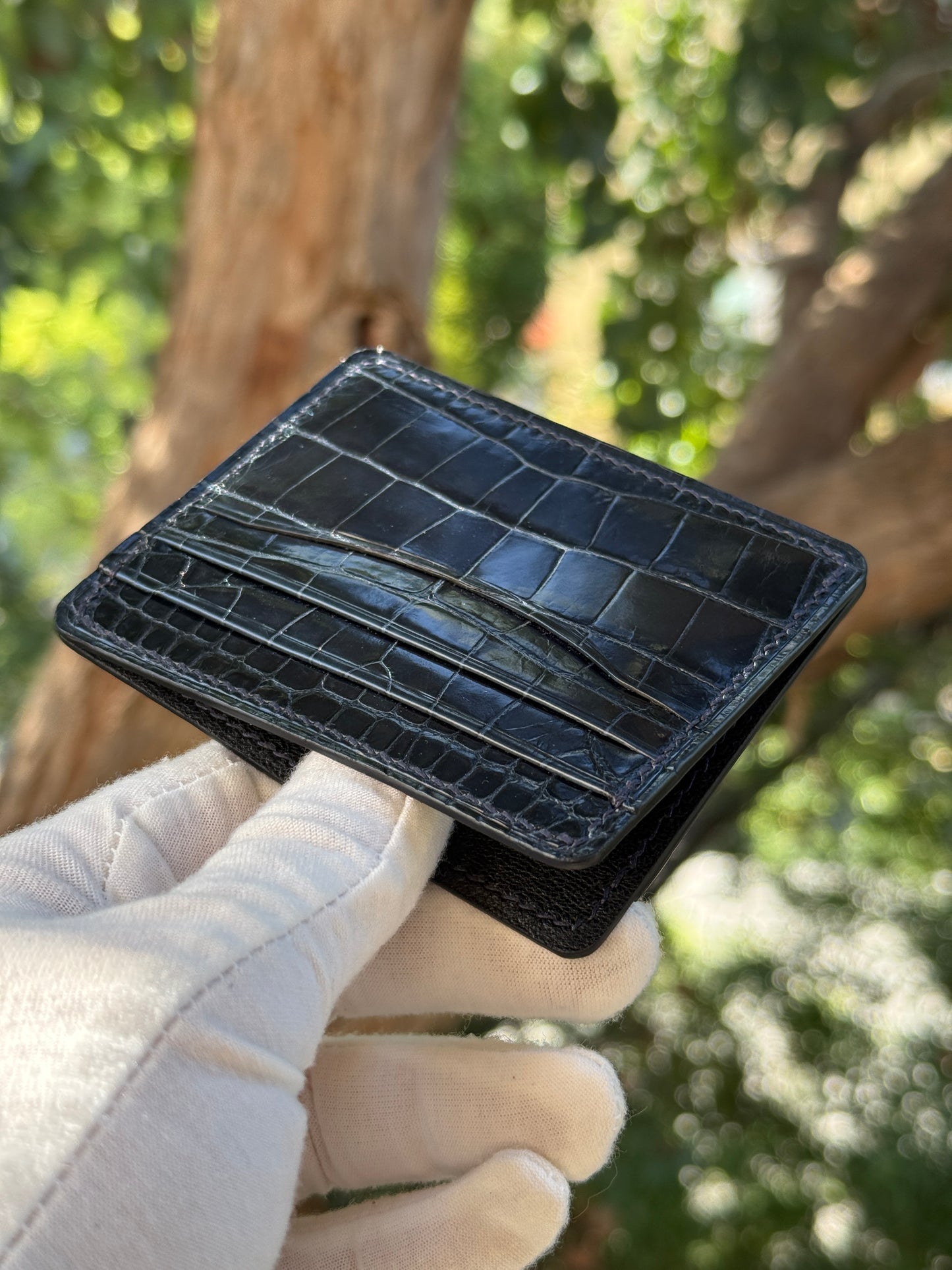 
                  
                    Navy glazed alligator leather card holder on a neutral background, showing its high-gloss scale texture and card slots.
                  
                