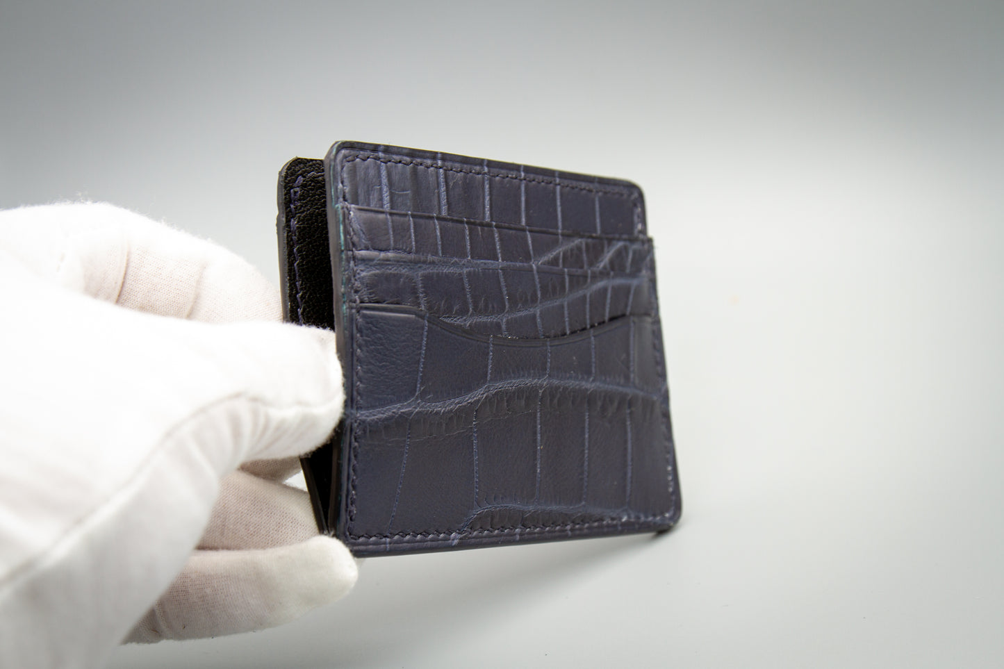 
                  
                    Another view of hand holding a matte navy alligator leather card case to show compact scale and real-world proportions.
                  
                