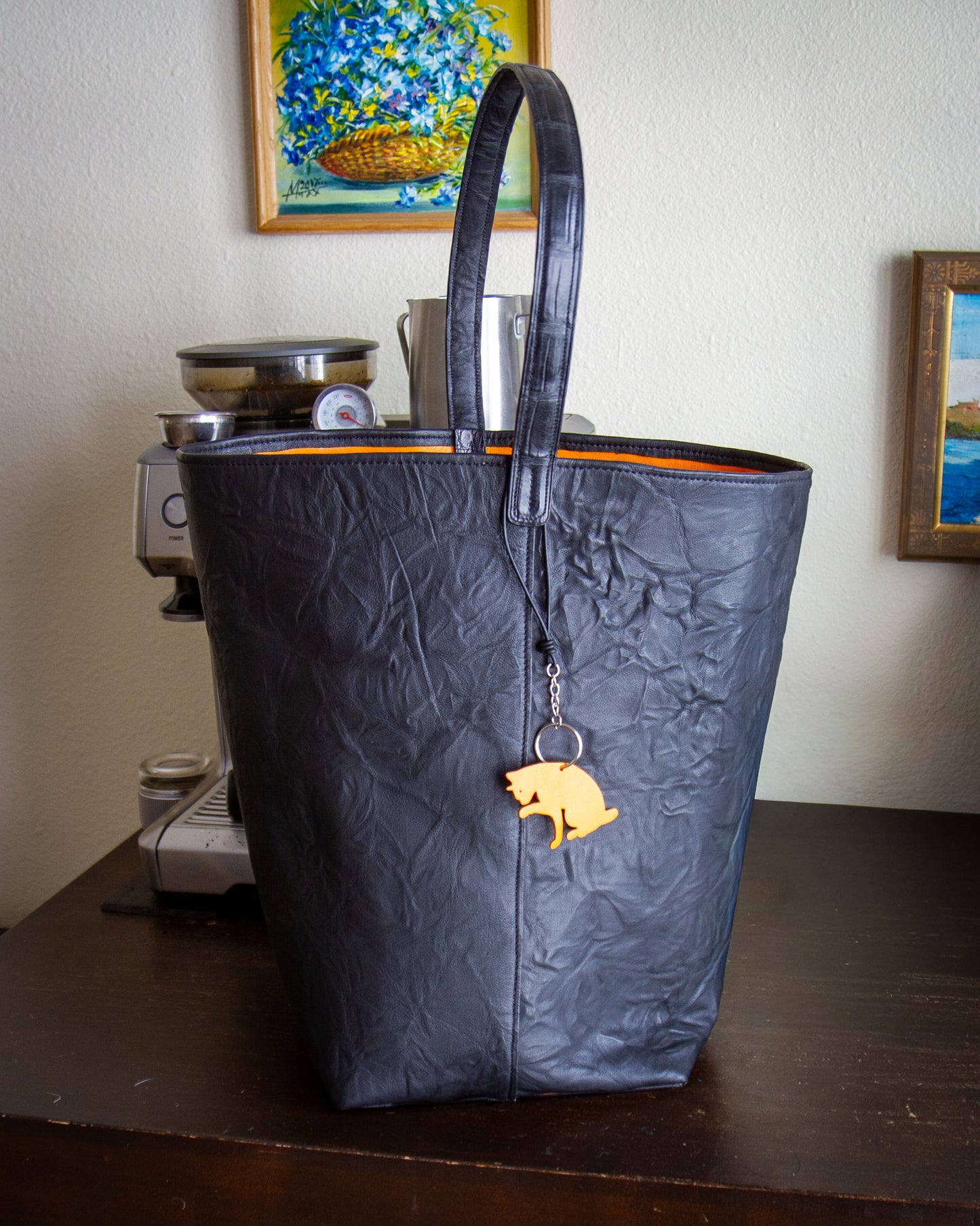 
                  
                    Angled close-up of the textured black goatskin exterior featuring a decorative orange cat charm attached by a leather cord
                  
                