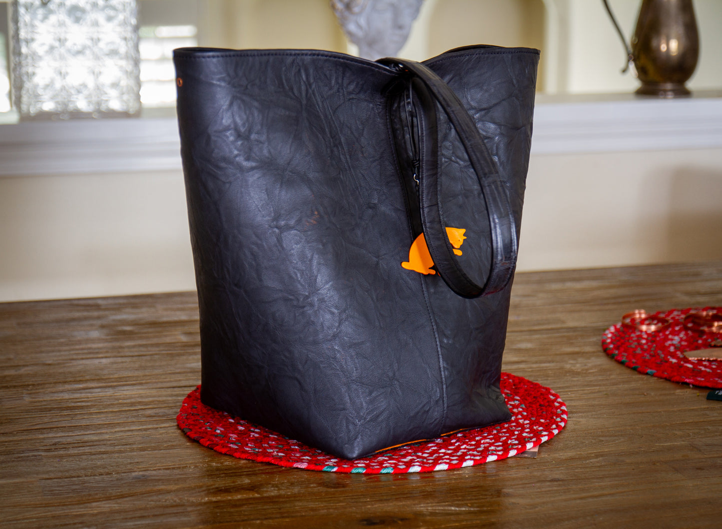 
                  
                    Front view of a tall black wrinkled goat leather tote bag with an embossed shoulder strap and hanging orange leather cat charm.
                  
                